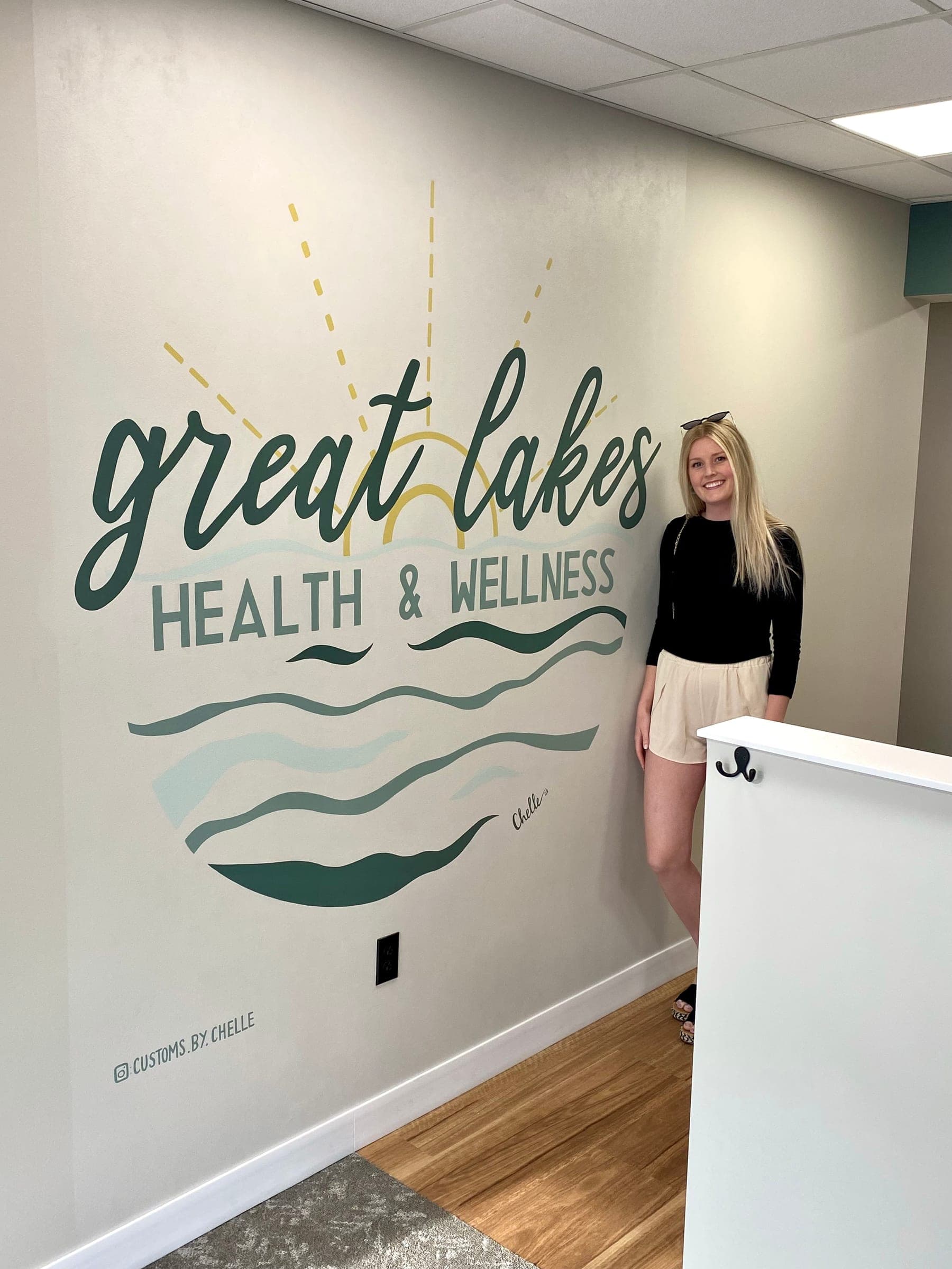 A woman standing beside the painted Great Lakes Health & Wellness sun and waves on an office wall