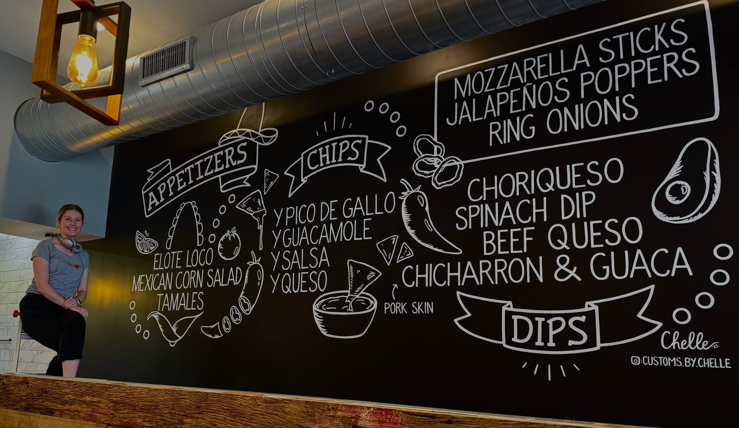 A long chalk-style menu wall lettered with appetizers, chips and dips, with a woman sitting at one end